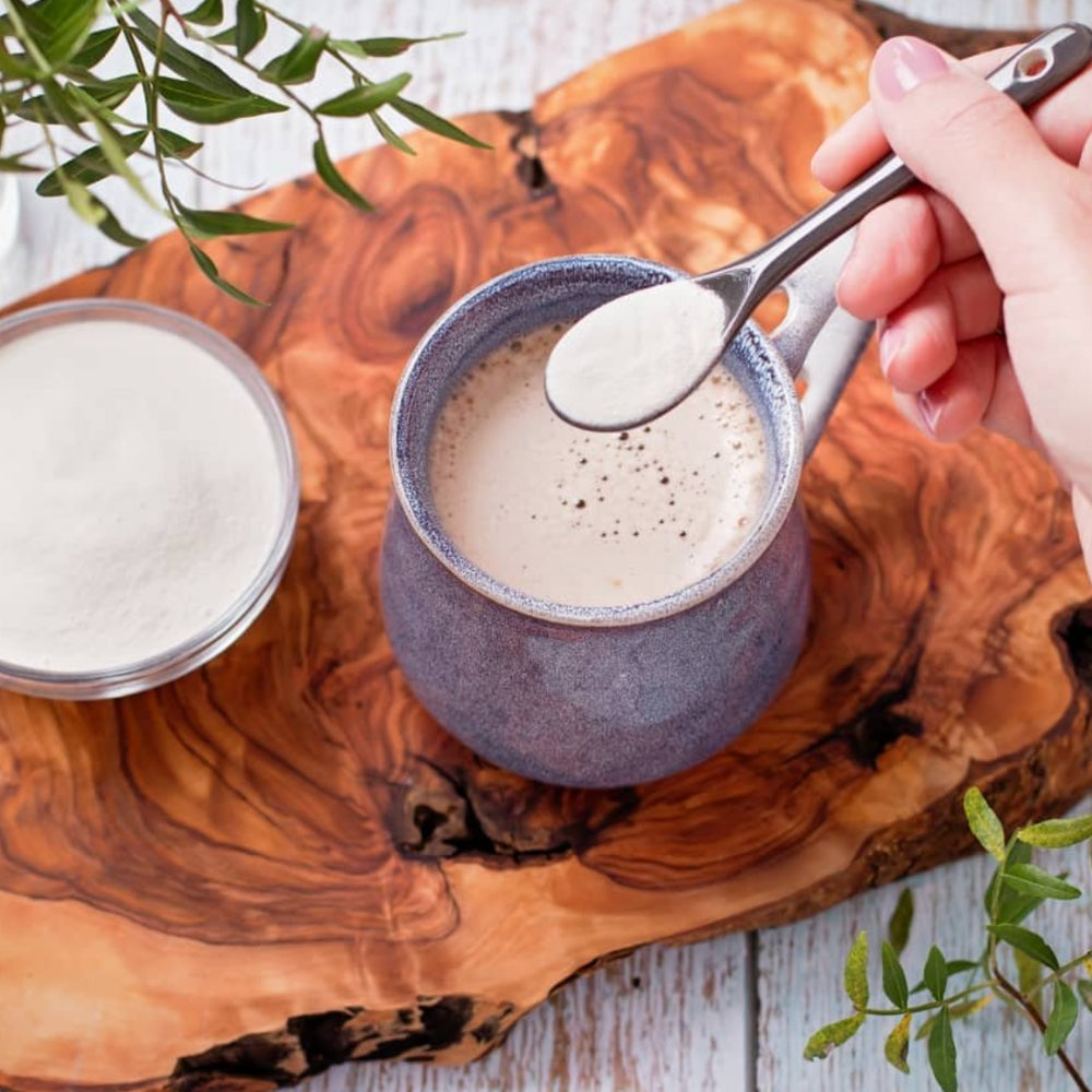Hand adding a spoonful of white powdered supplement to a frothy latte in a purple ceramic mug on a wood board